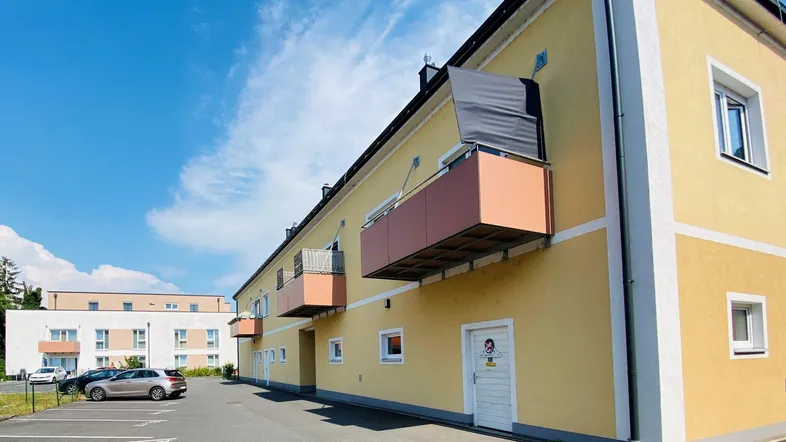 Gelbe Fassade eines Mehrfamilienhauses mit Balkonen und Parkplätzen unter blauem Himmel.