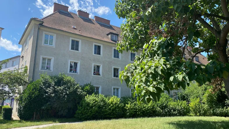 Ansicht eines Mehrfamilienhauses mit grüner Umgebung und Bäumen unter blauem Himmel.