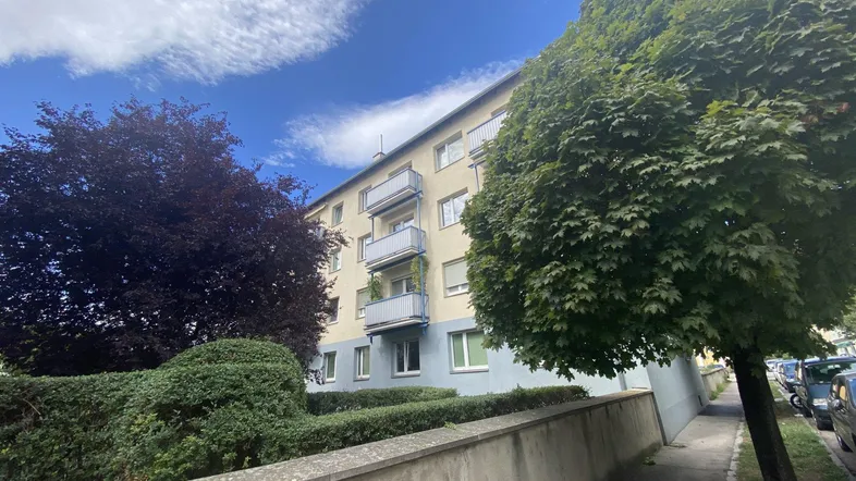 Mehrstöckiges Wohngebäude mit Balkonen und Bäumen unter blauem Himmel.