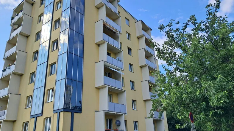 Modernes Mehrfamilienhaus mit gelber Fassade, Glasaufzug und zahlreichen Balkonen unter blauem Himmel.