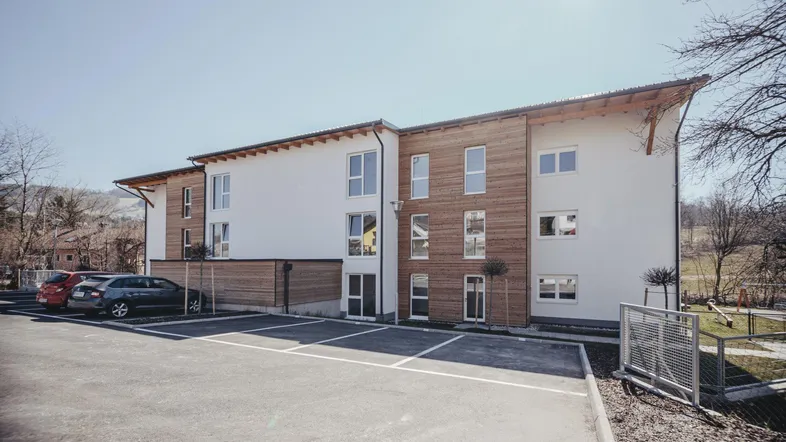 Modernes Mehrfamilienhaus mit Holzfassade und Parkplätzen unter strahlend blauem Himmel.