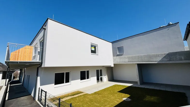 Modernes Neubaugebäude mit weißer Fassade, kleinem Gartenbereich und einem Balkon unter blauem Himmel.