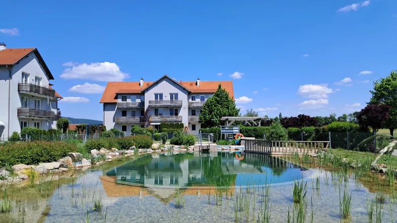 Großer Naturschwimmteich vor den Wohngebäuden, umgeben von grüner Landschaft unter blauem Himmel.