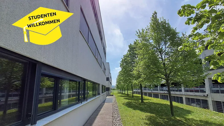Modernes Gebäude mit großen Fenstern und einem grünen Weg, der von Bäumen gesäumt ist. Ein Studenten-Willkommensschild ist sichtbar.