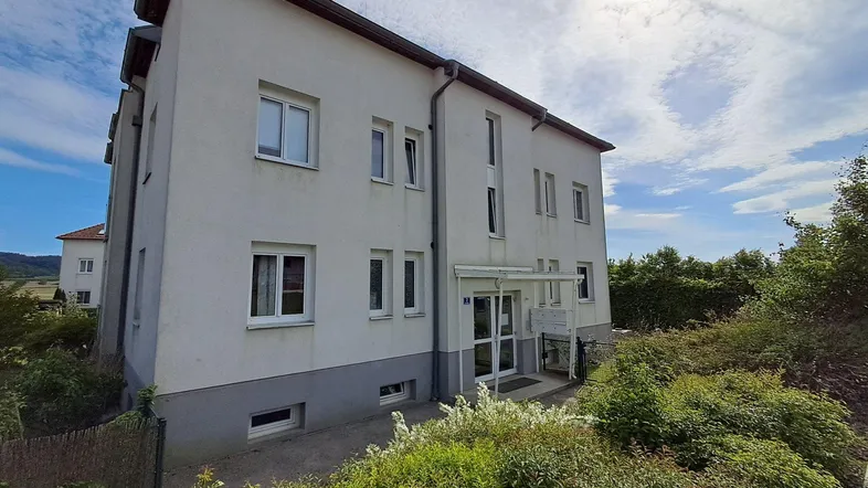 Weißes Mehrfamilienhaus mit grauer Sockelzone und umliegender Grünfläche unter blauem Himmel.