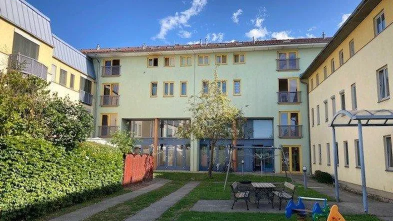 Außenansicht des modernen Wohngebäudes mit begrüntem Innenhof, Kinderspielplatz und Sitzgelegenheiten.