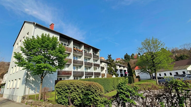 Mehrfamilienhaus mit mehreren Balkonen und grüner Umgebung unter blauem Himmel.