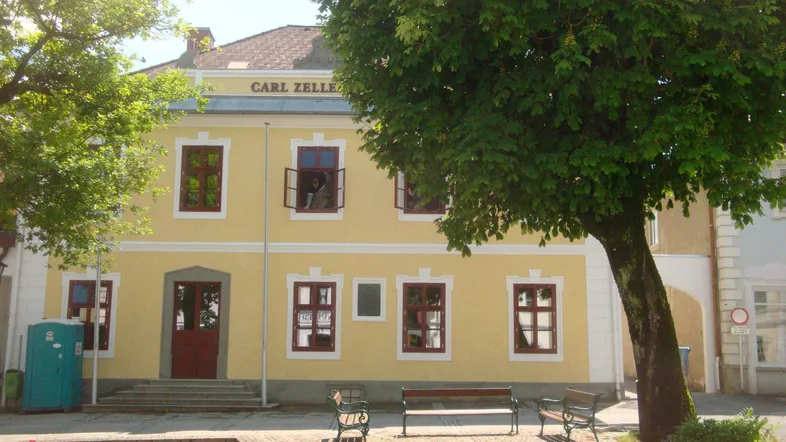 Historisches gelbes Gebäude mit traditionellen Fenstern und Bäumen im Vordergrund an einem sonnigen Tag.