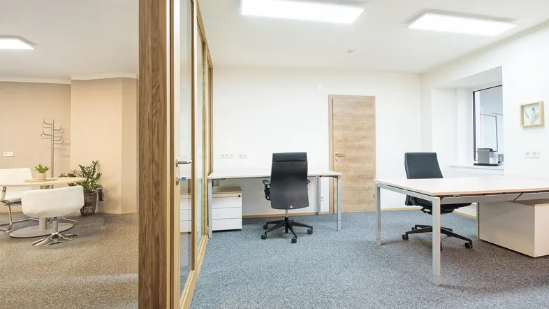 Helles Büro mit modernen Schreibtischen, Bürostühlen und Teppichboden, getrennt durch eine Glaswand.