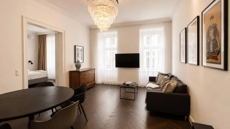 Heller Wohn- und Essbereich mit elegantem Kronleuchter, dunklem Fischgrätparkett und Zugang zum Schlafzimmer.