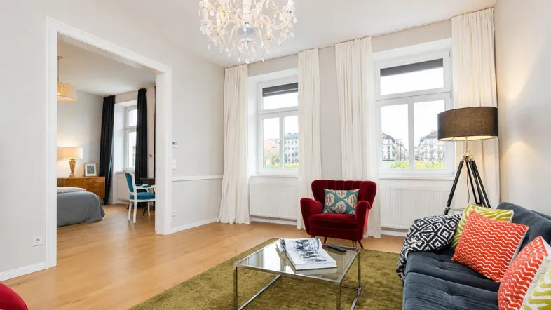 Helles Wohnzimmer mit elegantem Kronleuchter, bequemen Sitzmöbeln und Blick in ein angrenzendes Schlafzimmer.