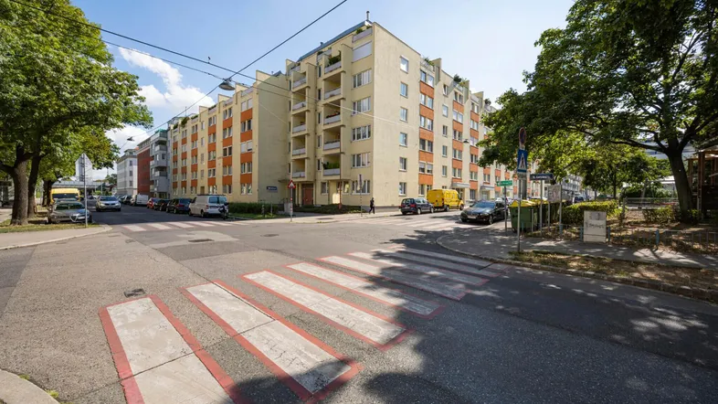 Mehrfamilienhaus an einer Straßenecke mit Bäumen, Zebrastreifen und geparkten Fahrzeugen.