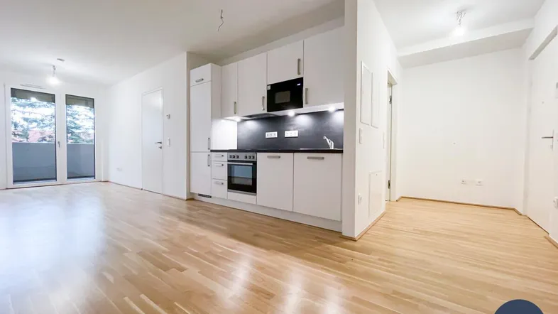 Heller offener Wohnbereich mit integrierter Küchenzeile, Parkettboden und Zugang zum Balkon.