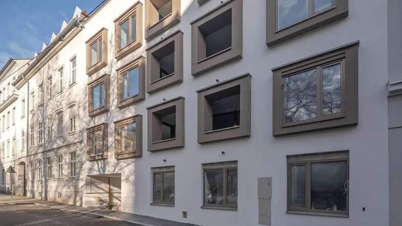 Moderne Fassade eines Wohngebäudes mit markanten Fenstern und hellem Putz in urbaner Umgebung.