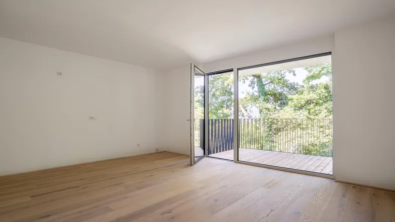 Helles Zimmer mit Holzboden und Zugang zu einem Balkon mit Blick ins Grüne.