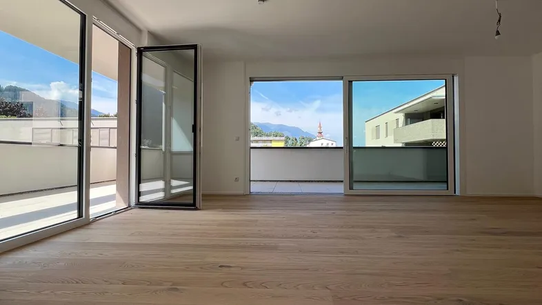 Heller, leerer Raum mit Eichenparkettboden, großen Fenstern und Zugang zu einem Balkon mit Bergblick.