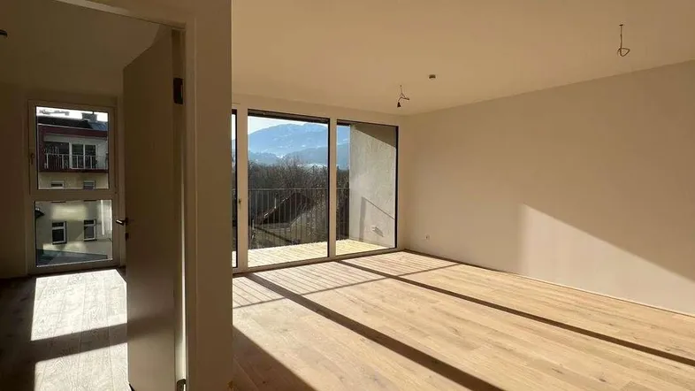 Helles, leeres Zimmer mit Holzboden und großen Schiebetüren, die zu einem Balkon mit Bergblick führen.