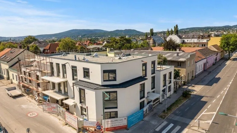 Luftaufnahme eines modernen Neubaus an einer Straßenecke mit umliegender Bebauung und grüner Landschaft im Hintergrund.
