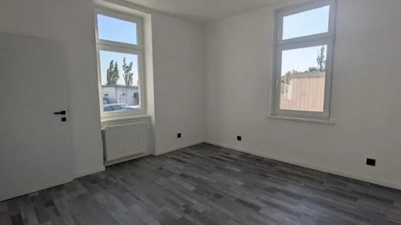 Helles, unmöbliertes Zimmer mit grauem Laminatboden und zwei Fenstern, bereit zur individuellen Gestaltung.