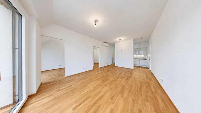Helles, leeres Zimmer mit Holzboden und Zugang zu einem Balkon, offener Bereich zur Küche.