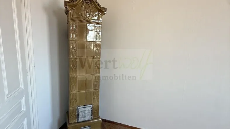 Helles Zimmer mit Fischgrätparkettboden und einem dekorativen Kachelofen in der Ecke.