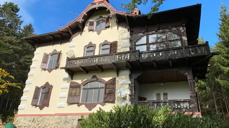 Historisches Gebäude mit aufwendiger Holzfassade, Balkonen und traditionellen Fensterläden in grüner Umgebung.