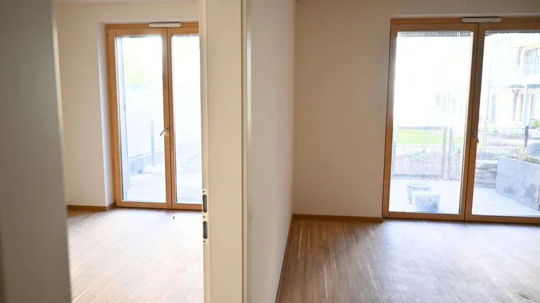 Heller Raum mit Holzboden und großen Fenstertüren, die Zugang zu einem Außenbereich bieten.