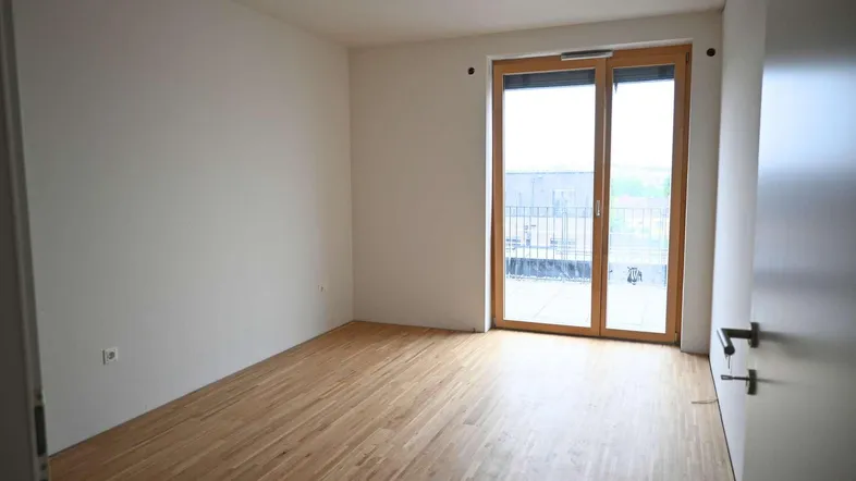 Helles, leeres Zimmer mit Holzboden und direktem Zugang zu einem Balkon.