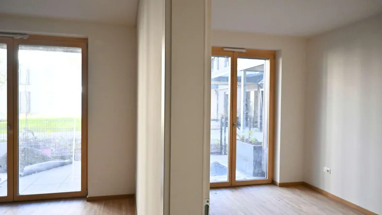 Helles, leeres Zimmer mit Holzboden und großen Fenstertüren, die ins Freie führen.