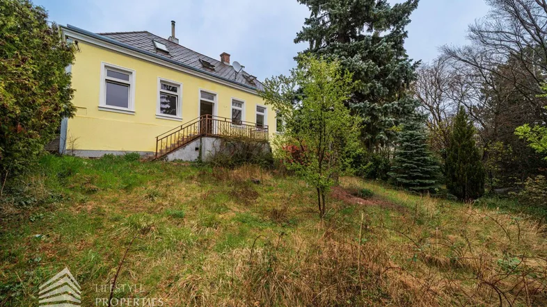 Gelbes Haus auf einem grasbewachsenen Hang mit hohen Bäumen unter bewölktem Himmel.