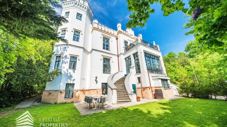 Elegantes weißes Schlossgebäude mit Turm, umgeben von üppigem Grün und einer sonnigen Terrasse.
