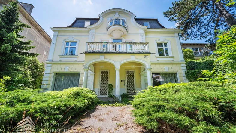 Historische Villa mit gelber Fassade, Balkon und üppiger Vegetation im Vorgarten.