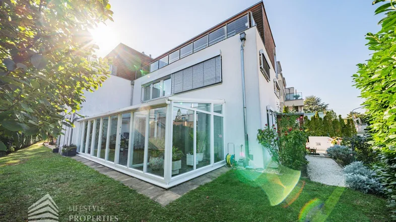 Modernes Haus mit Wintergarten und gepflegtem Garten unter strahlendem Sonnenschein.