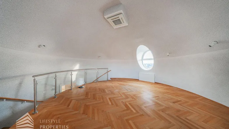 Heller Raum mit geschwungener Wand, Fischgrätparkett und einem runden Fenster, das viel Licht hereinlässt.