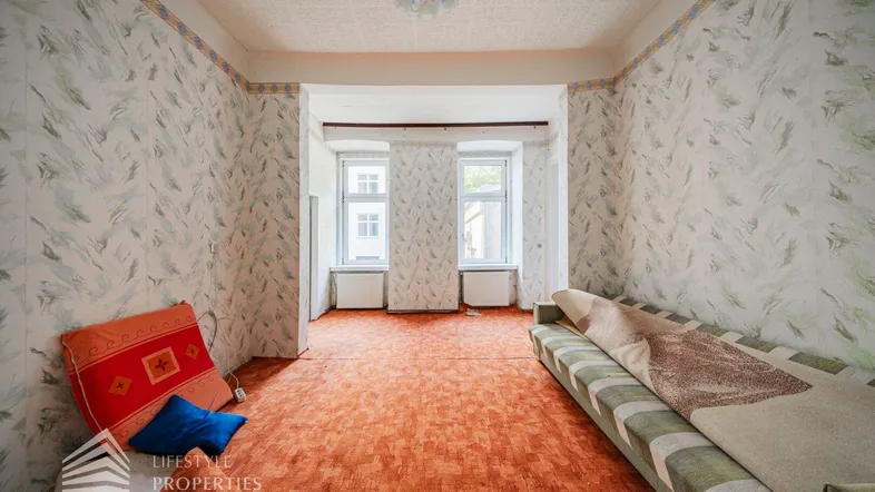 Geräumiges Zimmer mit gemusterter Tapete, Teppichboden und Blick nach draußen, renovierungsbedürftig.