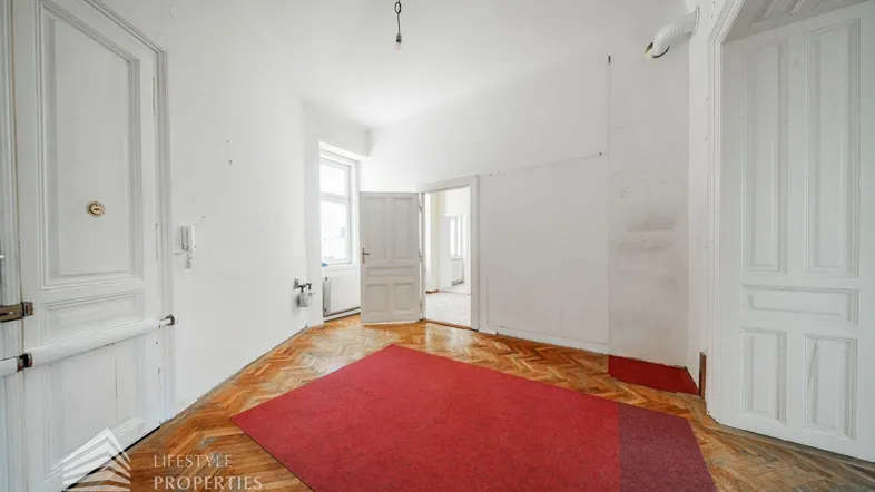 Leerer Raum mit Fischgrätparkett, hohen weißen Türen und einem roten Teppich in der Mitte.