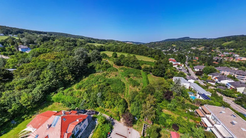 Luftaufnahme eines Wohngebiets mit viel Grün, Hügeln und einem Haus mit rotem Dach.