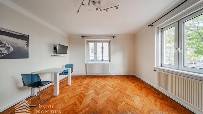 Helles Zimmer mit Fischgrätparkett, zwei Fenstern und einem kleinen Esstisch, ideal als Wohnbereich.