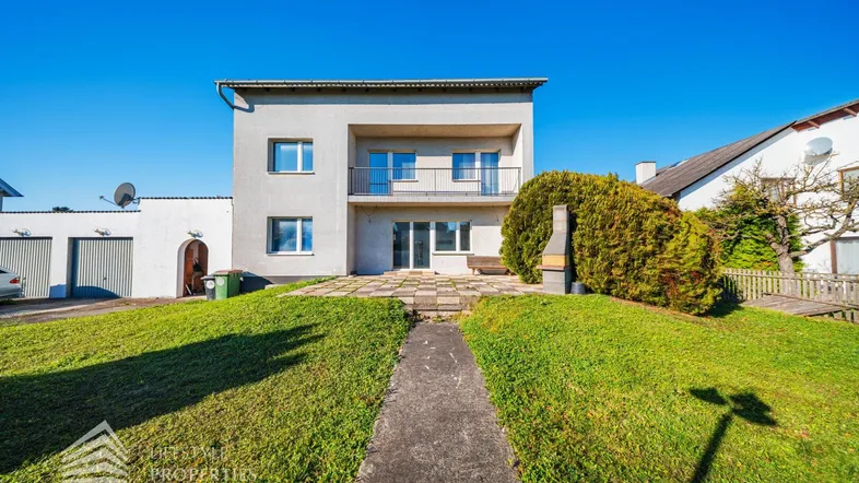 Vorderansicht des Hauses mit Balkon, Rasenfläche und Zufahrt zu den Garagen.