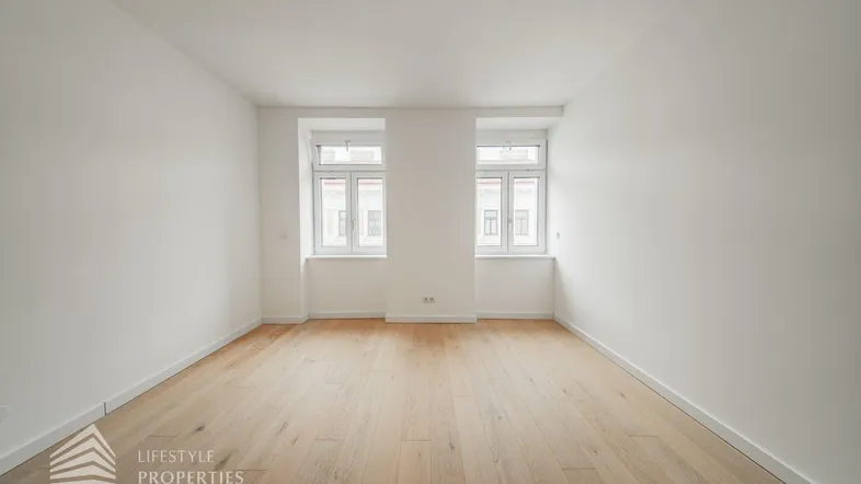 Helles, leeres Zimmer mit Parkettboden und zwei Fenstern, bereit zur individuellen Gestaltung.