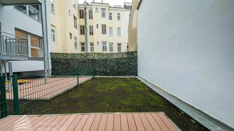 Kleiner Gartenbereich mit Holzterrasse und Hecke im Innenhof, umgeben von Gebäuden.