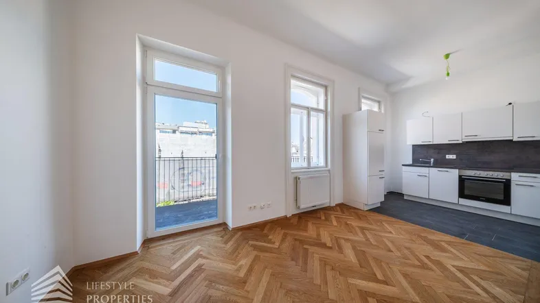 Heller offener Wohnbereich mit Fischgrätparkett und integrierter Küchenzeile sowie Zugang zu einem französischen Balkon.