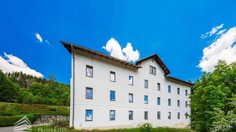 Weißes Gebäude mit grünen Fensterrahmen und Satteldach, umgeben von üppiger Natur und blauem Himmel.