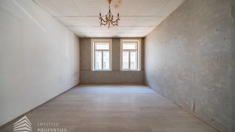 Geräumiger, unmöblierter Raum mit zwei Fenstern, Kronleuchter und Renovierungsbedarf.