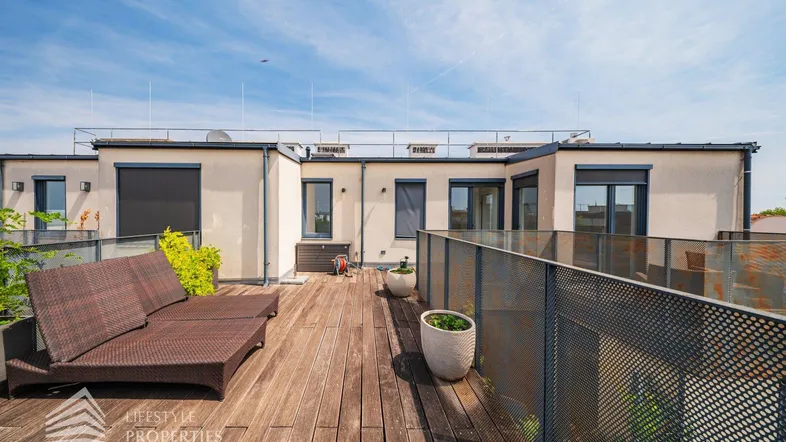 Geräumige Dachterrasse mit Holzdeck, Sonnenliege und Blick auf die Umgebung unter blauem Himmel.