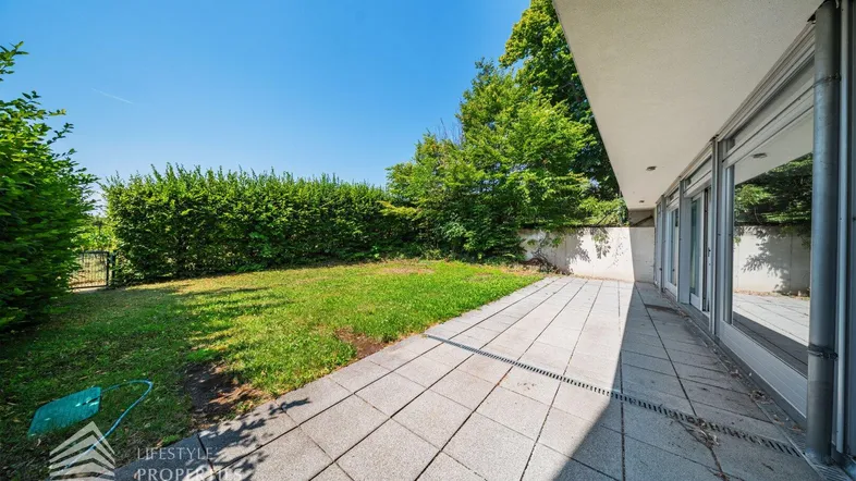 Geräumige Terrasse mit Steinplatten und angrenzendem grünen Rasenbereich, umgeben von einer hohen Hecke.