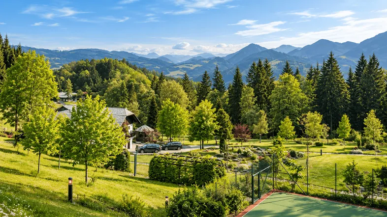 Weitläufige Gartenanlage mit Tennisplatz und malerischem Bergblick unter blauem Himmel.