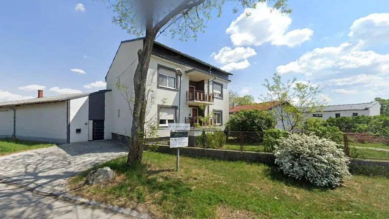 Hausfassade mit angrenzendem Gebäude und Zufahrt unter blauem Himmel.