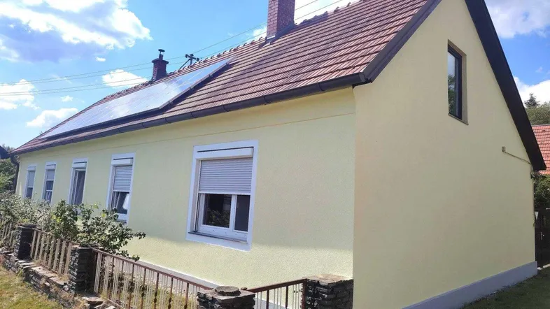 Gelb gestrichenes Haus mit Ziegeldach und Rollläden unter blauem Himmel.