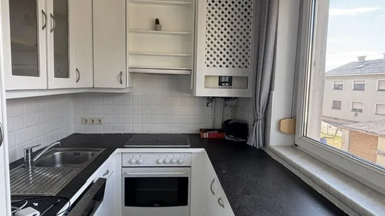 Kleine Küche mit weißen Schränken, dunkler Arbeitsplatte und Blick nach draußen durch das Fenster.